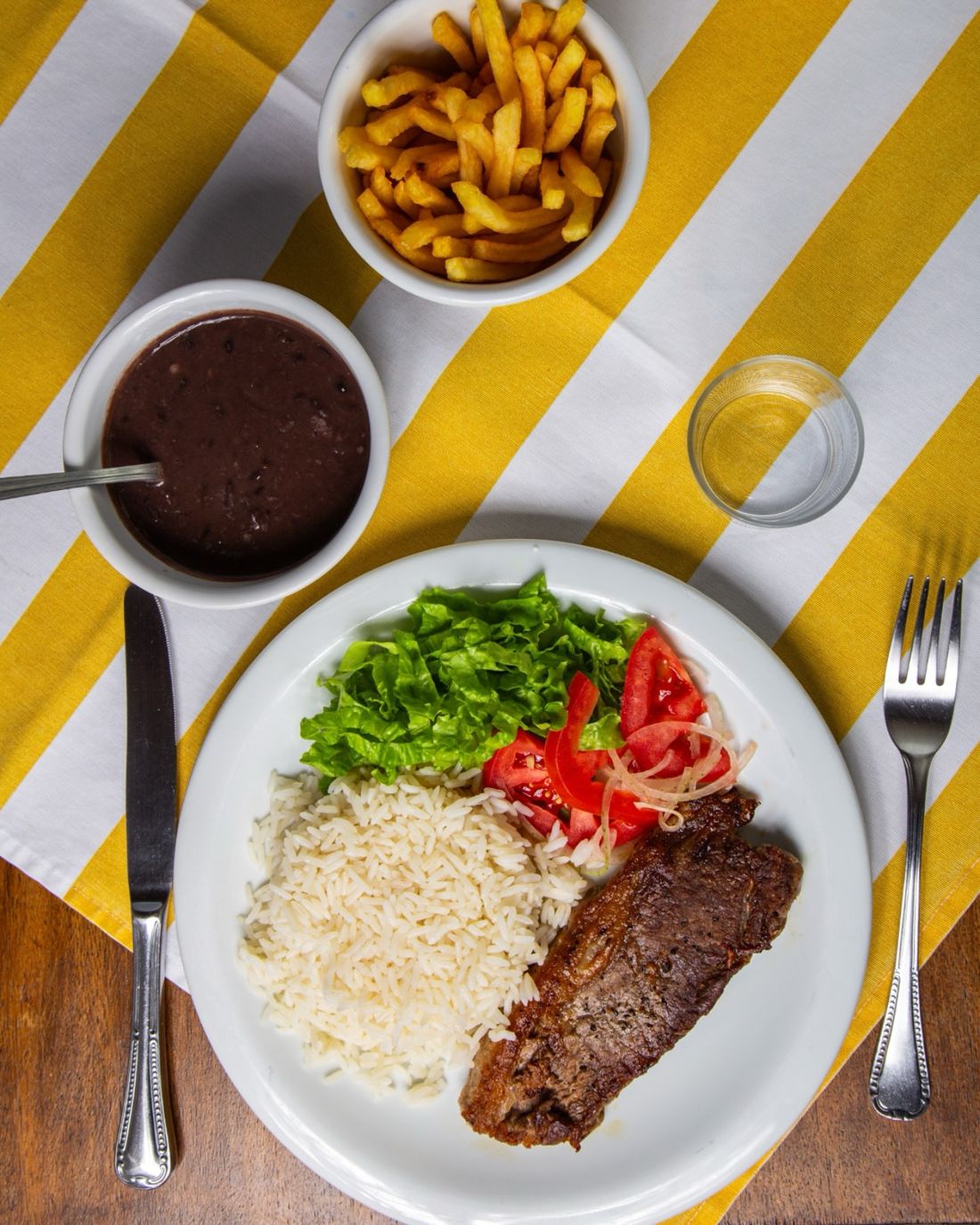 Prato executivo de carne vermelha com arroz, salada, feijão e batata frita na toalha listrada amarela do By Cuca