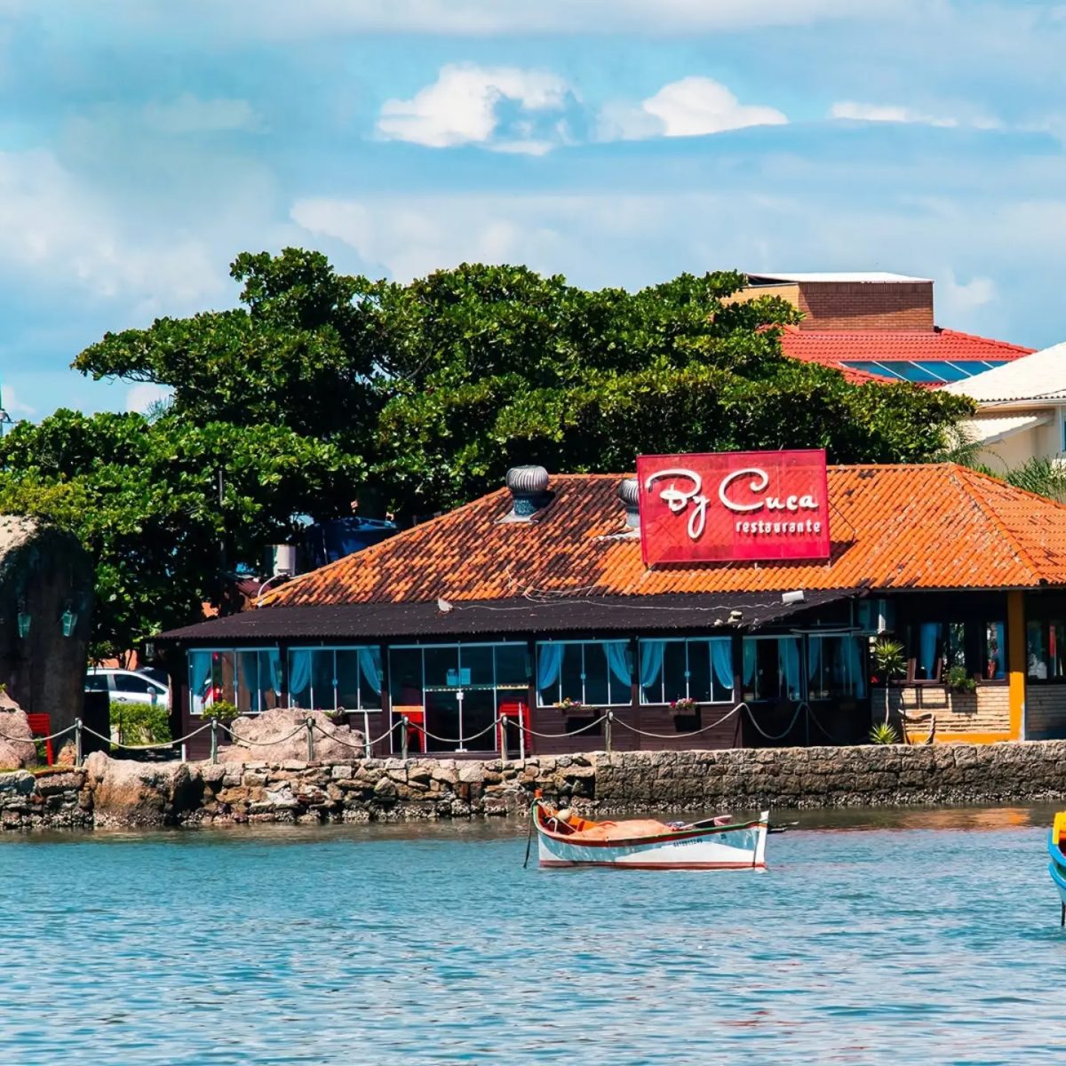Fachada do By Cuca Restaurante à beira-mar, com barco de pesca ancorado em frente