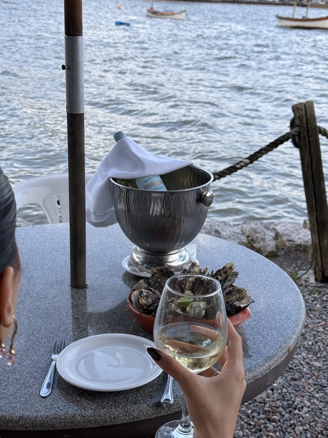 Taça de vinho branco erguida diante do mar, com ostras e balde de gelo na mesa à beira-mar do By Cuca